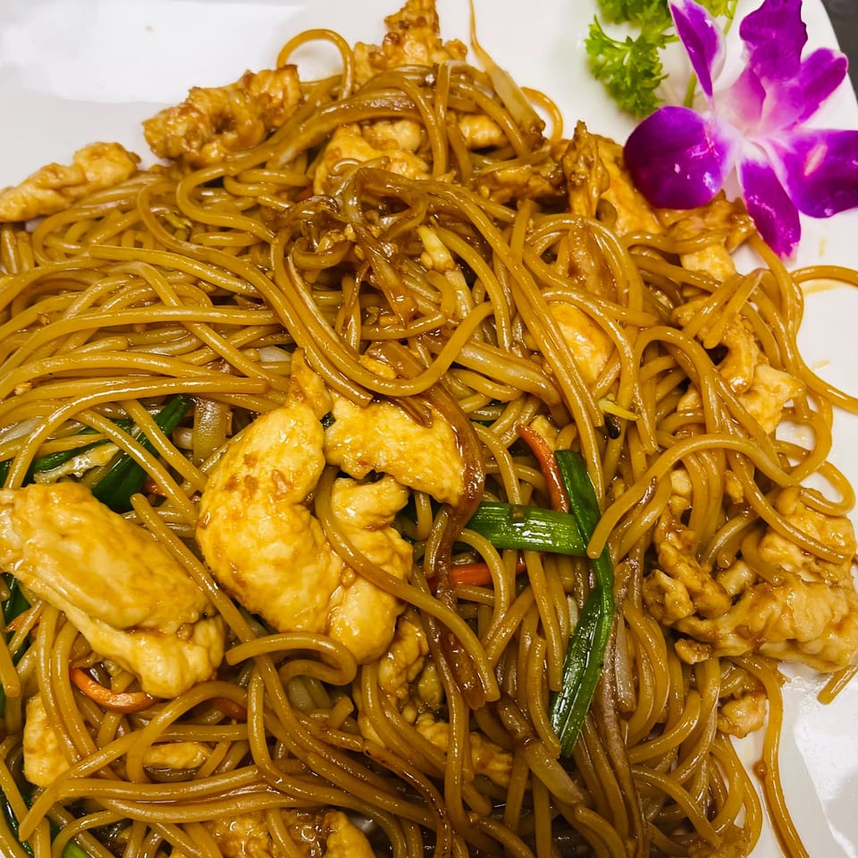 Close-up of Thai chicken noodles with green onions and a purple orchid garnish.