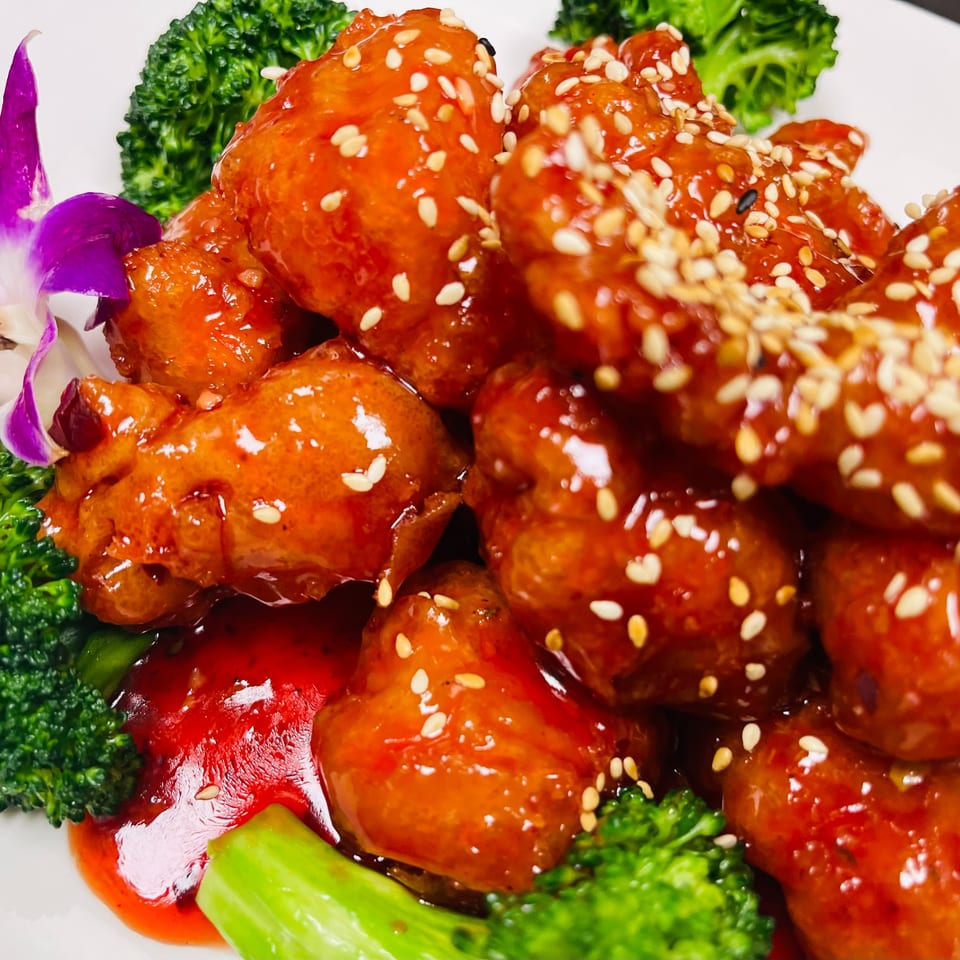 Orange chicken with sesame seeds, broccoli, and a purple orchid on a white plate.