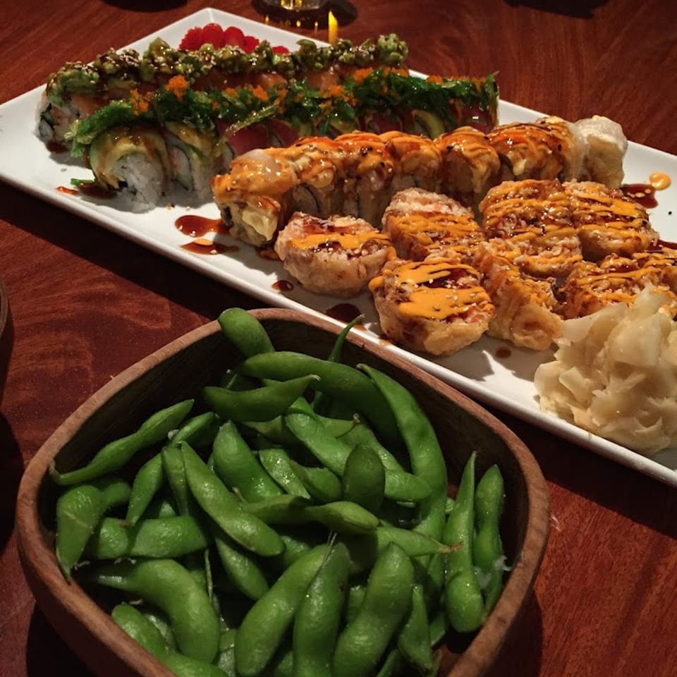 A plate of sushi rolls with various toppings and a bowl of edamame on a wooden table.